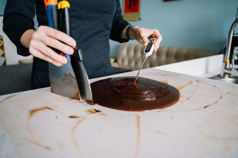 Close-up of a person tempering chocolate on a kitchen counter using a scraper and thermometer.